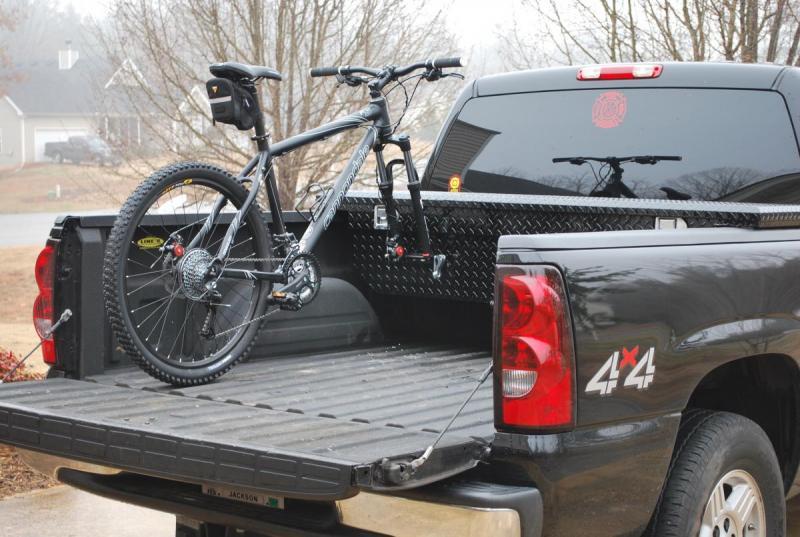 Cannondale F5: A black mountain bike secured in the bed of a pickup truck, which has a diamond plate toolbox and a "4x4" sticker on its side. The background features a foggy residential area with trees and homes. The scene conveys an outdoor and adventure theme.