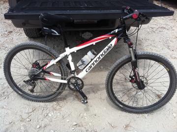 Cannondale F7: A Cannondale mountain bike with a white and red frame, placed on a dirt surface beside a black truck. The bike features front suspension, disc brakes, and a water bottle holder.