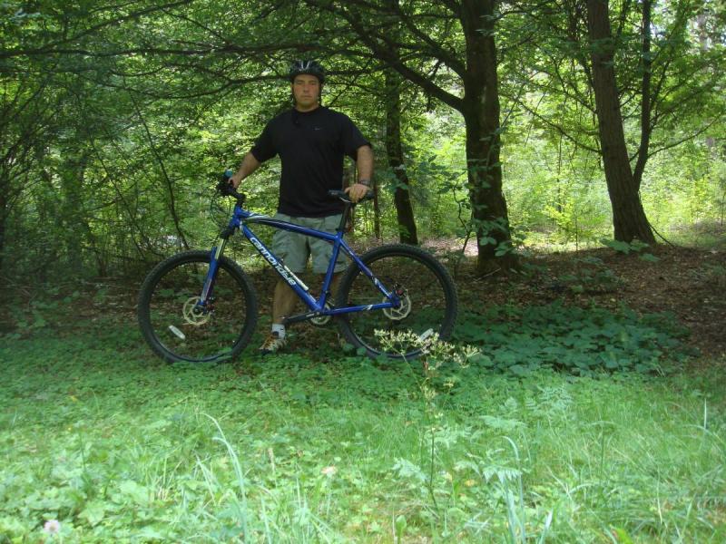 Cannondale F7: A person wearing a black t-shirt and helmet stands beside a blue mountain bike in a wooded area. The ground is covered with grass and foliage, and trees surround the scene, providing a lush green backdrop. The atmosphere suggests an outdoor adventure or cycling activity.