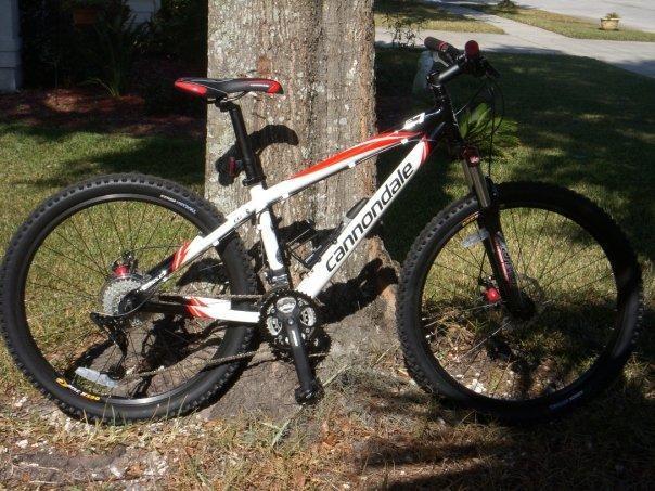 Cannondale F7: A mountain bike parked next to a tree, featuring a white and red Cannondale frame, black tires, and prominent suspension. The scene is set in a grassy area, with a glimpse of a driveway in the background.