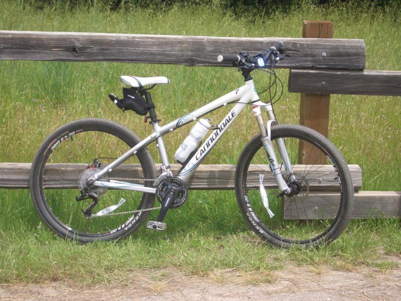 Cannondale F7: A white Cannondale mountain bike leaning against a wooden fence, with green grass in the background. The bike features knobby tires, a water bottle holder, and is positioned on a gravel path, suggesting a recreational outdoor setting.