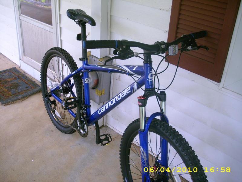 Cannondale F7: A blue mountain bike leaning against a porch, featuring knobby tires and a sturdy frame. The bike has a suspension fork and is positioned next to a front door with a welcome mat visible on the ground.