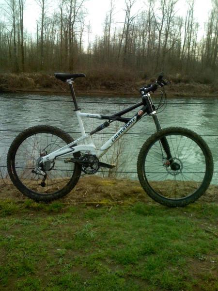 Cannondale Prophet 5: A mountain bike with a black and white frame is positioned on a grassy area beside a river. Trees are visible in the background, indicating a natural setting. The scene suggests an outdoor recreational environment suitable for biking activities.