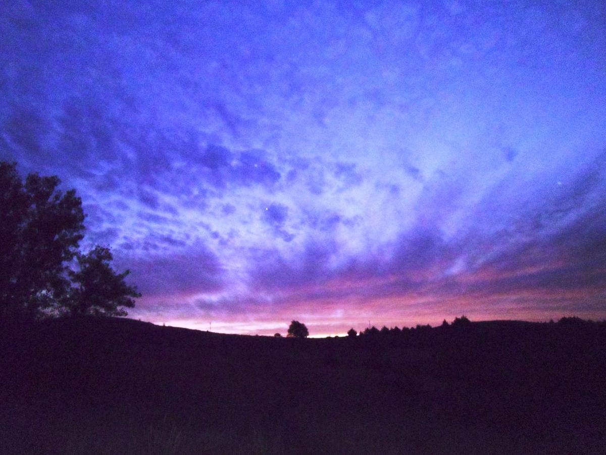 A dramatic evening sky displaying shades of purple and blue, with wispy clouds scattered across. Silhouettes of trees and hills provide a striking contrast against the colorful backdrop. Switchgrass mountain bike trail.