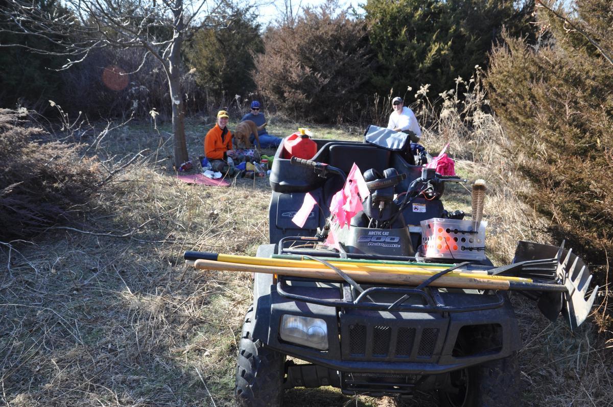 An all-terrain vehicle is parked in the foreground, loaded with tools and equipment. In the background, a small group of people sits on a picnic blanket in a grassy area. Trees and shrubs surround the location, indicating a natural outdoor setting. The scene captures a peaceful day outdoors with friends and nature. Switchgrass mountain bike trail.