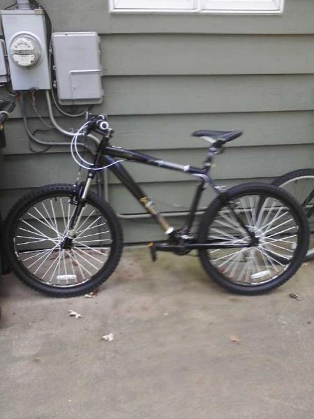 Diamondback Response: A black mountain bike is parked against a gray wall, with electrical equipment visible in the background. The bike features thick tires, a sturdy frame, and a slightly angled handlebar.