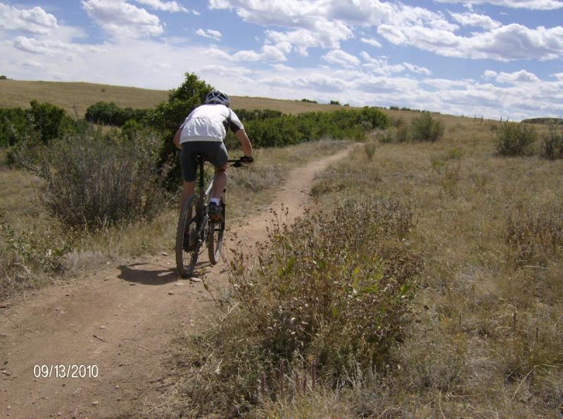 Diamondback Sortie 1: A person riding a mountain bike on a dirt path in a grassy landscape, with sparse bushes and a partly cloudy sky in the background.