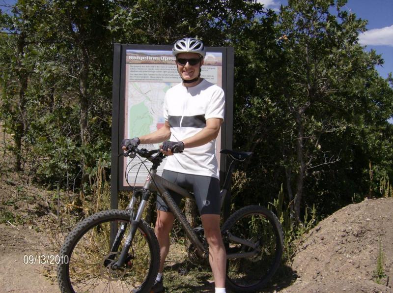 Diamondback Sortie 1: A person in a cycling helmet and sunglasses stands next to a mountain bike, smiling while holding the handlebars. They are dressed in a white and black cycling shirt and shorts. Behind them is an information sign, likely about the cycling area, with a map and text. The background features greenery and dirt, suggesting an outdoor location suitable for biking. The date on the image is September 13, 2010.