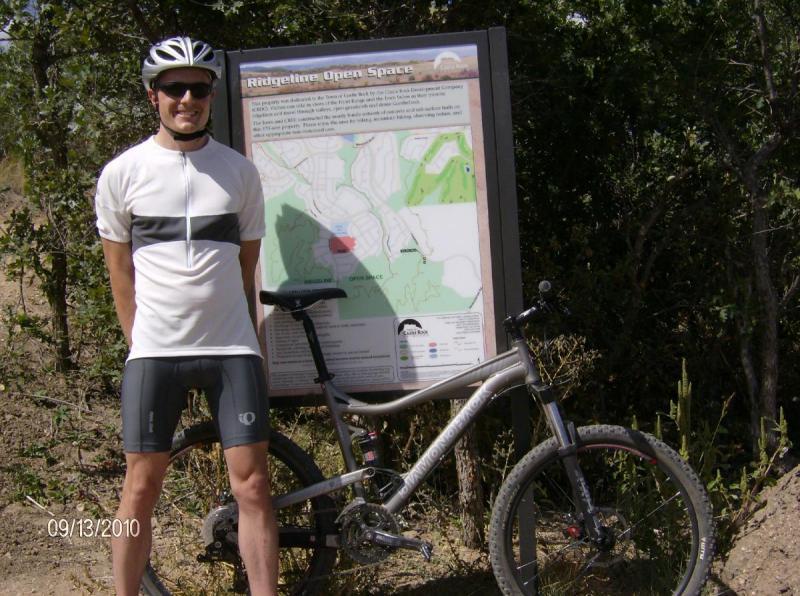 Diamondback Sortie 1: A cyclist wearing a helmet and athletic attire stands next to a mountain bike in front of a trail map sign for Ridgeline Open Space. The background features foliage and the map displays the area's trails and features.