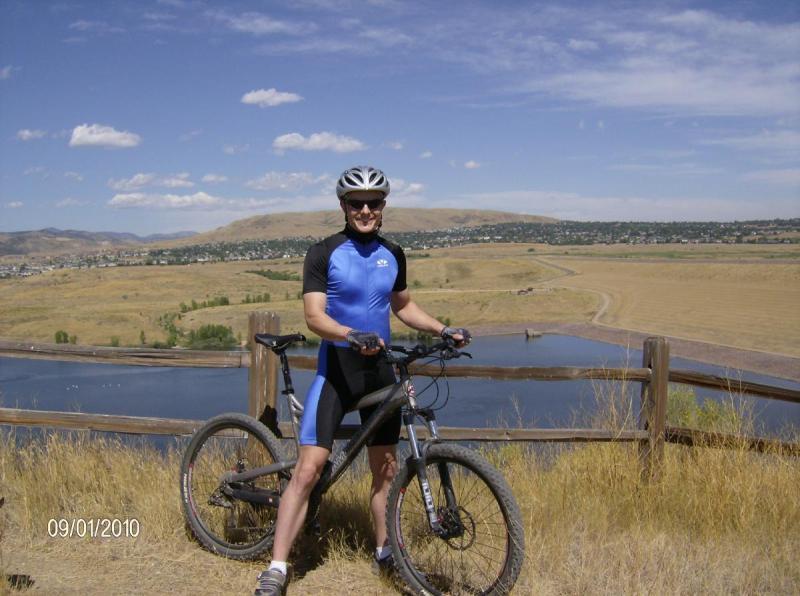 Diamondback Sortie 1: A cyclist in a blue and black biking outfit stands next to a mountain bike, smiling at the camera. The background features a scenic landscape with rolling hills, a body of water, and a clear blue sky with a few clouds.