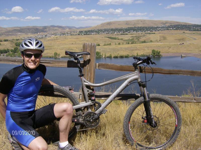 Diamondback Sortie 1: A person wearing a blue cycling jersey and helmet smiles while sitting next to a mountain bike, with a scenic landscape of hills, a lake, and blue skies in the background.