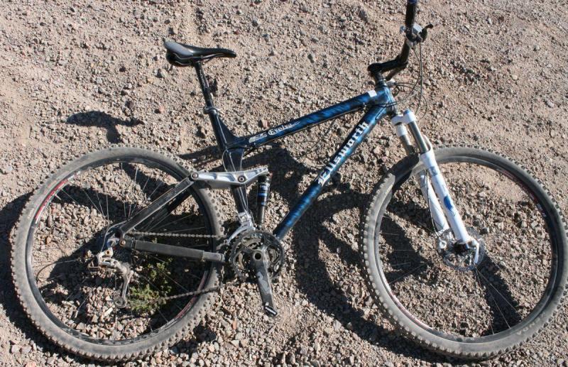 Ellsworth Evolve: A mountain bike lying on rocky terrain, featuring a blue and black frame, front suspension, and knobby tires. The bike shows signs of use, with dirt and gravel surrounding it.
