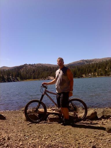 Ellsworth Moment: A person stands next to a mountain bike by a lake, surrounded by trees and mountains under a clear blue sky. They are wearing a sleeveless shirt and shorts, and the scenery reflects a serene outdoor environment.