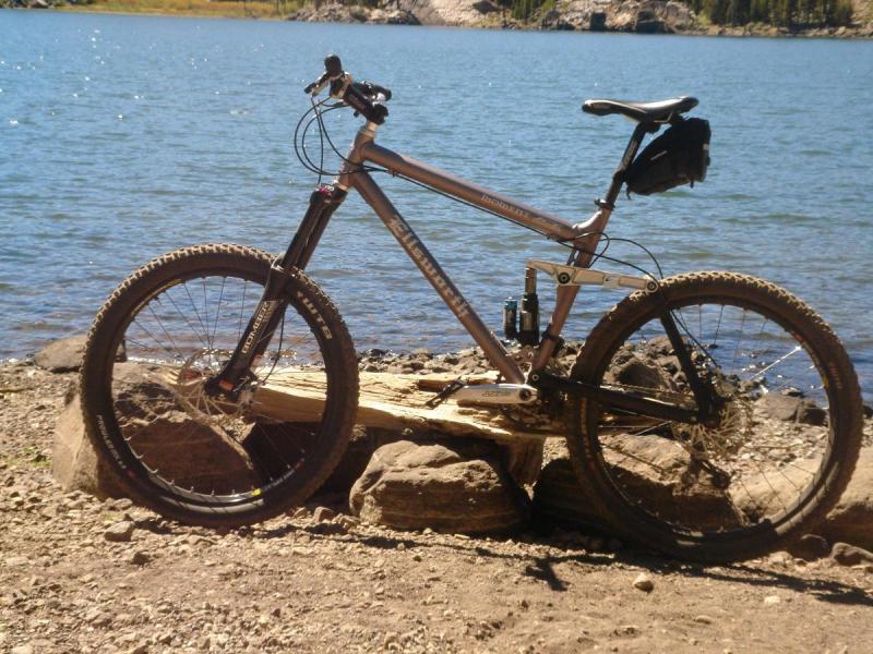 Ellsworth Moment: A mountain bike resting on large rocks by the edge of a clear blue lake, surrounded by trees and rugged terrain. The bike has a metallic frame and thick tires, designed for off-road use. Sunlight reflects off the water, creating a scenic outdoor setting.
