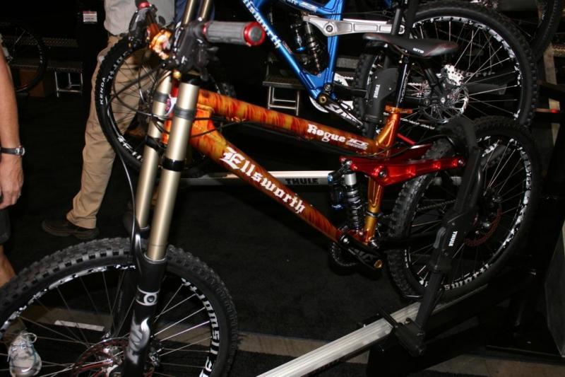 Ellsworth Rogue: Image of a close-up view of two mountain bikes displayed at an event. The foreground features a brightly colored Ellsworth bike with a gradient design and visible suspension components. In the background, a blue bike is partially obscured. Below, the bikes rest on a stand against a dark background, and a person is partially visible to the left, showcasing the setting.
