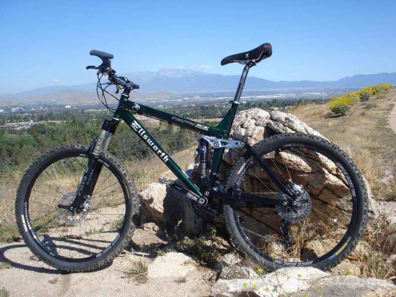 Ellsworth Truth: Mountain bike leaning against a rocky outcrop with a scenic landscape of hills and valleys in the background under a clear blue sky.