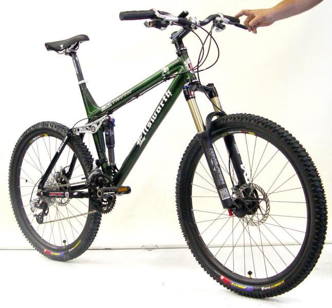 Ellsworth Truth: A green mountain bike with a sleek design, featuring a dual suspension system, wide tires, and disc brakes. The bike is positioned upright against a white background, with a hand reaching towards the handlebars.