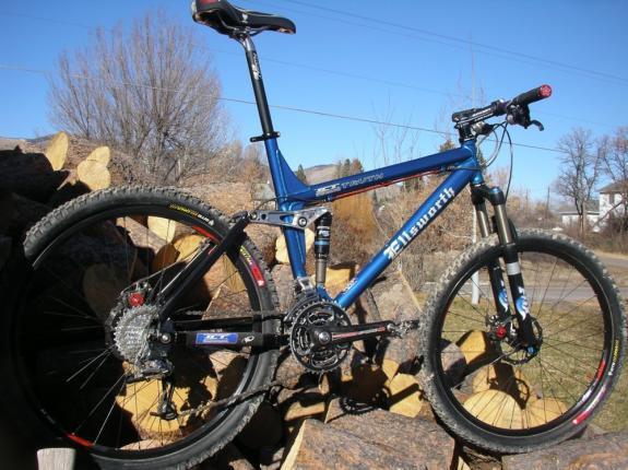 Ellsworth Truth: A blue mountain bike with a sturdy frame and suspension resting on a pile of logs. The bike features thick tires, a visible gear system, and a modern design, set against a clear blue sky and sparse trees in the background.
