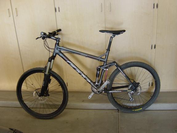 Felt Virtue Two: A black and silver mountain bike leaning against a wall in a garage setting, featuring thick tires and a front suspension fork, with a simple wooden cabinet in the background.