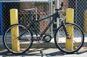 Fetish Fixation: A black mountain bike leaning against two yellow posts, set against a chain-link fence. The bike features thick tires, a gear shift, and a sleek design. Sunlight casts shadows on the ground.