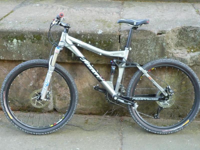 Fezzari Nebo Peak: A silver mountain bike with a black frame, featuring dual suspension and thick tires, leaning against a moss-covered stone wall. The bike has a visible disc brake on the front wheel and colorful accents on the wheels.