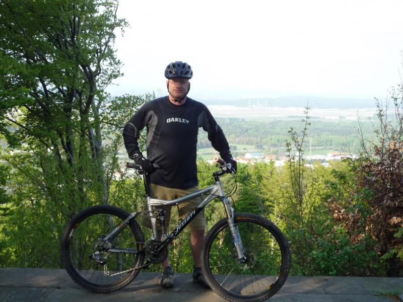 Fezzari Nebo Peak: A man in a black long-sleeve shirt and helmet stands beside a silver mountain bike on a scenic overlook, surrounded by greenery and trees. In the background, a panoramic view of a valley is visible, with distant hills and structures.