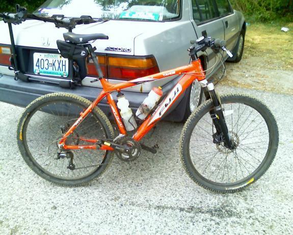 Fuji Tahoe Pro: An orange mountain bike parked next to a gray car. The bike has a water bottle attached to the frame and features knobby tires suitable for off-road riding. The background includes greenery and a gravel surface.
