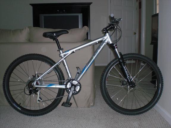 GT Avalanche: A silver mountain bike with black tires and a lightweight frame, positioned in a living room setting. The bike is standing next to a beige couch, with a television visible in the background.