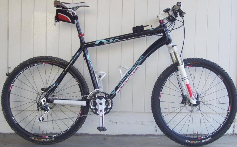 Gary Fisher Big Sur: Mountain bike featuring a black and white frame with aqua accents, knobby tires, front suspension, and a dual chainring setup, positioned against a white wooden wall.