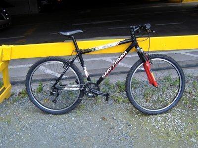 Gary Fisher Big Sur: A black mountain bike with red front suspension, parked next to a yellow barrier on a gravel surface. The bike features wide tires and a gear system, indicating it's designed for off-road use.