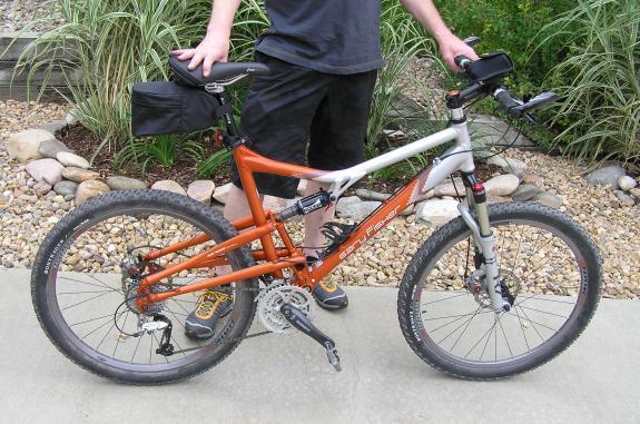 Gary Fisher Caliber: A person stands next to an orange and silver mountain bike on a concrete path, surrounded by decorative stones and greenery. The bike features thick tires and a small black bag attached to the frame. The individual is wearing a black T-shirt and shorts, with their hand resting on the bike's handlebar.
