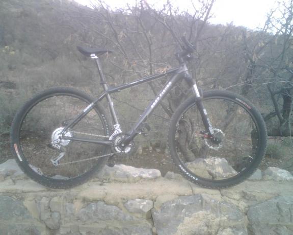 Gary Fisher Cobia: A mountain bike is resting on a stone wall with rugged terrain in the background. The bike features a black frame, thick tires, and front suspension, indicating it's designed for off-road use. Surrounding the bike are sparse bushes and rocky ground, suggesting a natural outdoor setting.