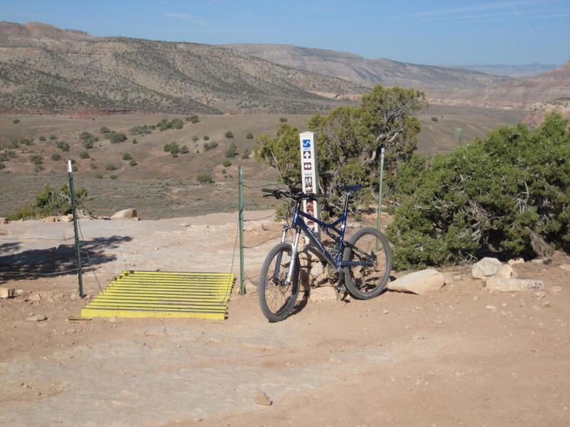 Gary Fisher HiFi Plus: A mountain bike rests against a post near a trail sign, with a metal grate in the foreground. The landscape features rolling hills and distant mountains under a clear blue sky.