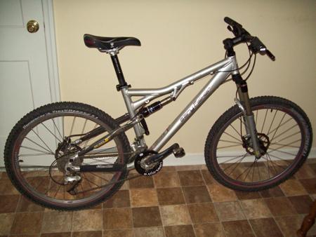 Gary Fisher HiFi Plus: A silver mountain bike with thick tires and a suspension system, leaning against a wall in a room with a tiled floor.