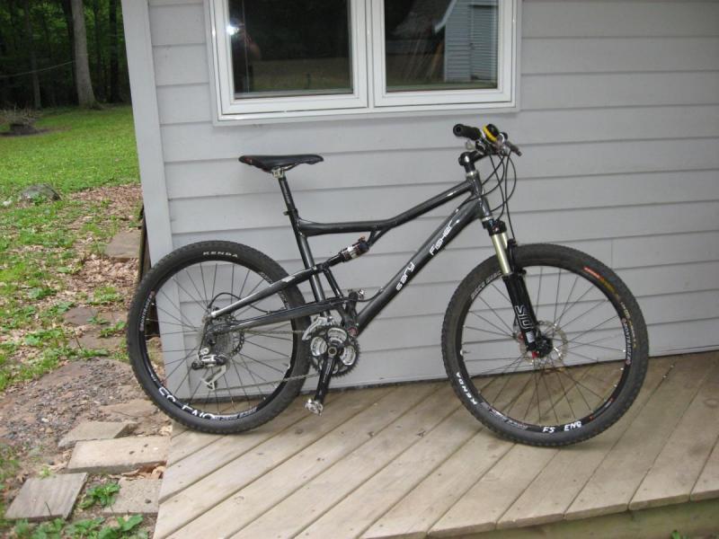 Gary Fisher HiFi Pro: A black mountain bike with a sturdy frame and thick tires is leaning against a wooden deck next to a gray house. The bike features a front suspension fork and multiple gears, with a scenic green lawn visible in the background.