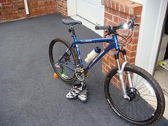 Gary Fisher Hoo Koo E Koo: A blue mountain bike with a water bottle attached, positioned on a gray carpet in an indoor setting. A pair of cycling shoes are placed next to the bike. The background features a brick wall and a white door.