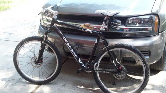 Gary Fisher Marlin Disc: A black mountain bike with a sleek design, featuring a front suspension and disc brakes, parked next to a gray SUV. The bike is positioned at an angle, showcasing its wheels and frame.