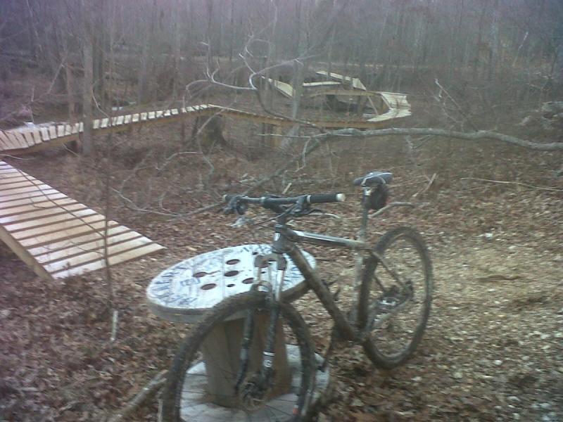 Gary Fisher Marlin: A mountain bike resting on a wooden platform in a wooded area with bare trees and a dirt trail visible. In the background, there are additional wooden structures and pathways, indicating a mountain biking trail system. The ground is covered with fallen leaves.