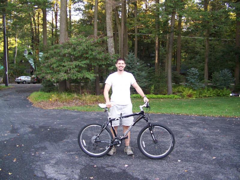 Gary Fisher Marlin: A young man stands on a paved driveway, smiling while holding a black mountain bike. He wears a white t-shirt and light-colored shorts. Surrounding him are tall trees and a well-maintained lawn with greenery in the background. A car is visible on the side of the driveway.