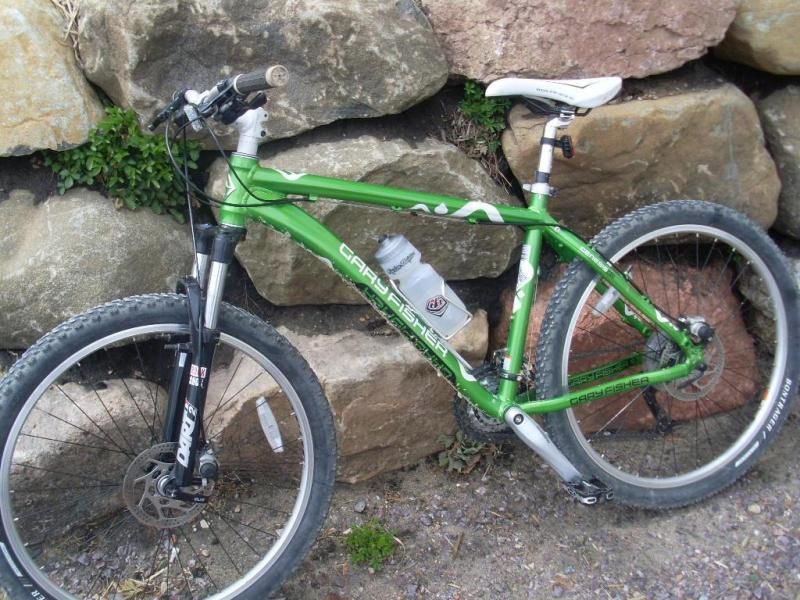 Gary Fisher Marlin: A green mountain bike leaning against a rocky wall, featuring thick tires and a water bottle attached to the frame. The bike displays a sturdy design, with visible components such as the handlebars and suspension. The surrounding area includes pebbles and greenery, suggesting an outdoor setting.