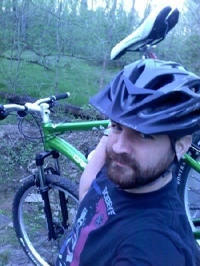 Gary Fisher Marlin: A person wearing a black helmet stands next to a green mountain bike in a wooded outdoor setting. The individual has a beard and is wearing a black t-shirt with a colorful graphic design. Trees and greenery are visible in the background, suggesting a natural environment suitable for biking.