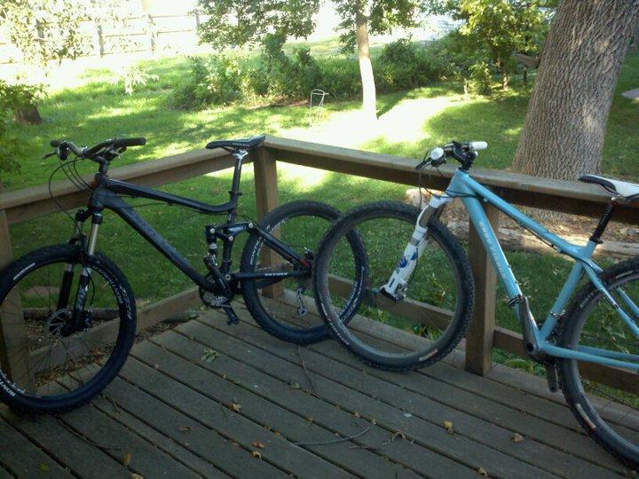 Gary Fisher Paragon: Two mountain bikes leaning against a wooden railing on a porch. The bike on the left is black, while the bike on the right is light blue. In the background, there is a grassy area with trees and a sunny outdoor setting.