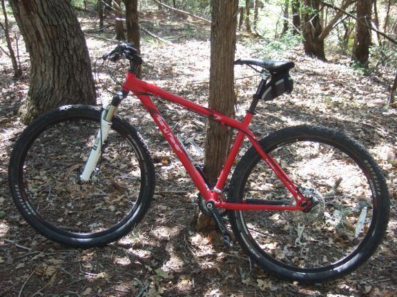 Gary Fisher Paragon: A red mountain bike with a front suspension fork, positioned among trees in a forested area. The bike features wide tires and a small bag attached to the seat, resting on a bed of leaves and dirt.
