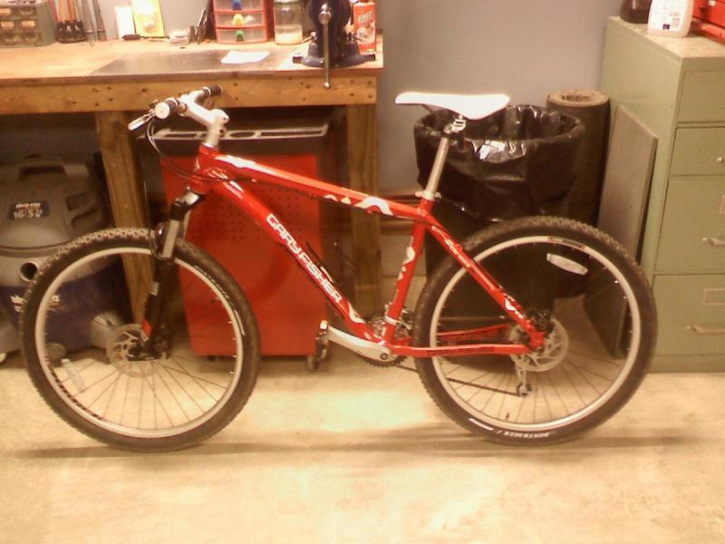 Gary Fisher Piranha: A red mountain bike with a white seat and black tires, positioned in a workshop setting with a wooden workbench in the background, a trash can, and various tools and materials.