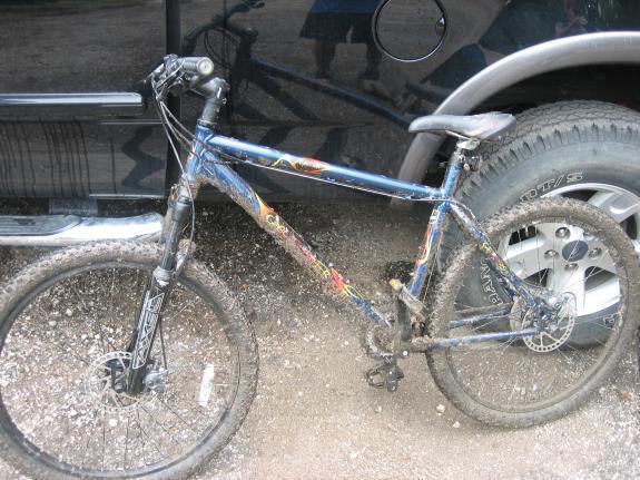 Gary Fisher Piranha: A mountain bike covered in mud, leaning against a dark vehicle. The bike features a blue frame with colorful graphics and has visible signs of wear and dirt. The background includes a large tire from the vehicle.
