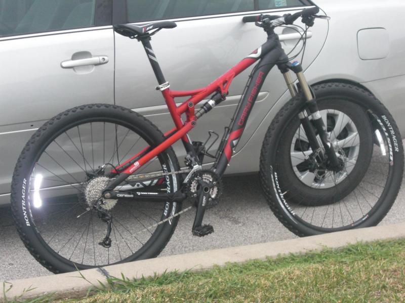 Trek Rumblefish: A black and red mountain bike parked beside a gray car, featuring wide tires and a dual suspension frame, with visible gear components and disc brakes. The bike is positioned on a concrete surface with grass along the edge.