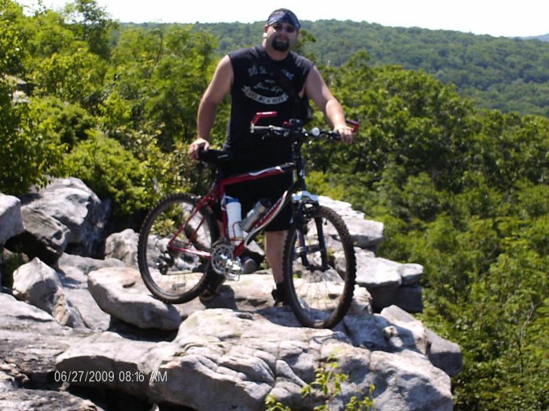 Gary Fisher Tarpon: A man stands on a rocky outcrop in a mountainous area, holding a mountain bike. He is wearing a sleeveless black shirt, shorts, and a bandana. Lush green trees and hills are visible in the background under clear blue skies.