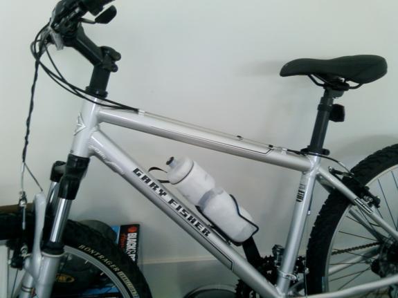 Gary Fisher Tarpon: A silver Gary Fisher mountain bike is shown, featuring a water bottle holder mounted on the frame. The bike has black handlebars and seat, and a thick tire visible in the background. It is positioned against a light-colored wall, and there are some items on the floor beneath it.