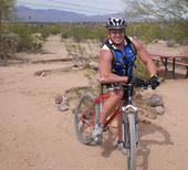 Gary Fisher Tassajara: A muscular cyclist wearing a helmet and a blue athletic vest, seated on a red mountain bike in a desert landscape with sparse vegetation and distant mountains.