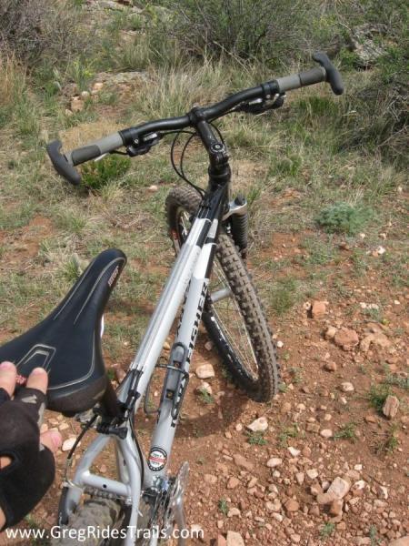 Gary Fisher Tassajara: Close-up view of a mountain bike on a rocky, dirt trail surrounded by grass and shrubs. The image shows the bike's handlebars and seat, with a focus on the knobby tires designed for off-road cycling.